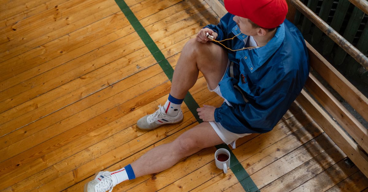 Sports coach in casual attire takes a break, sitting on the gym's wooden floor with a whistle.